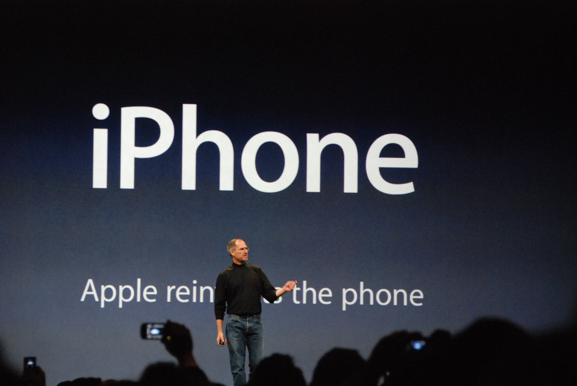 Steve Jobs standing in front of slide at the January 2007 iPhone introduction showing the tagline "Apple reinvents the phone"
