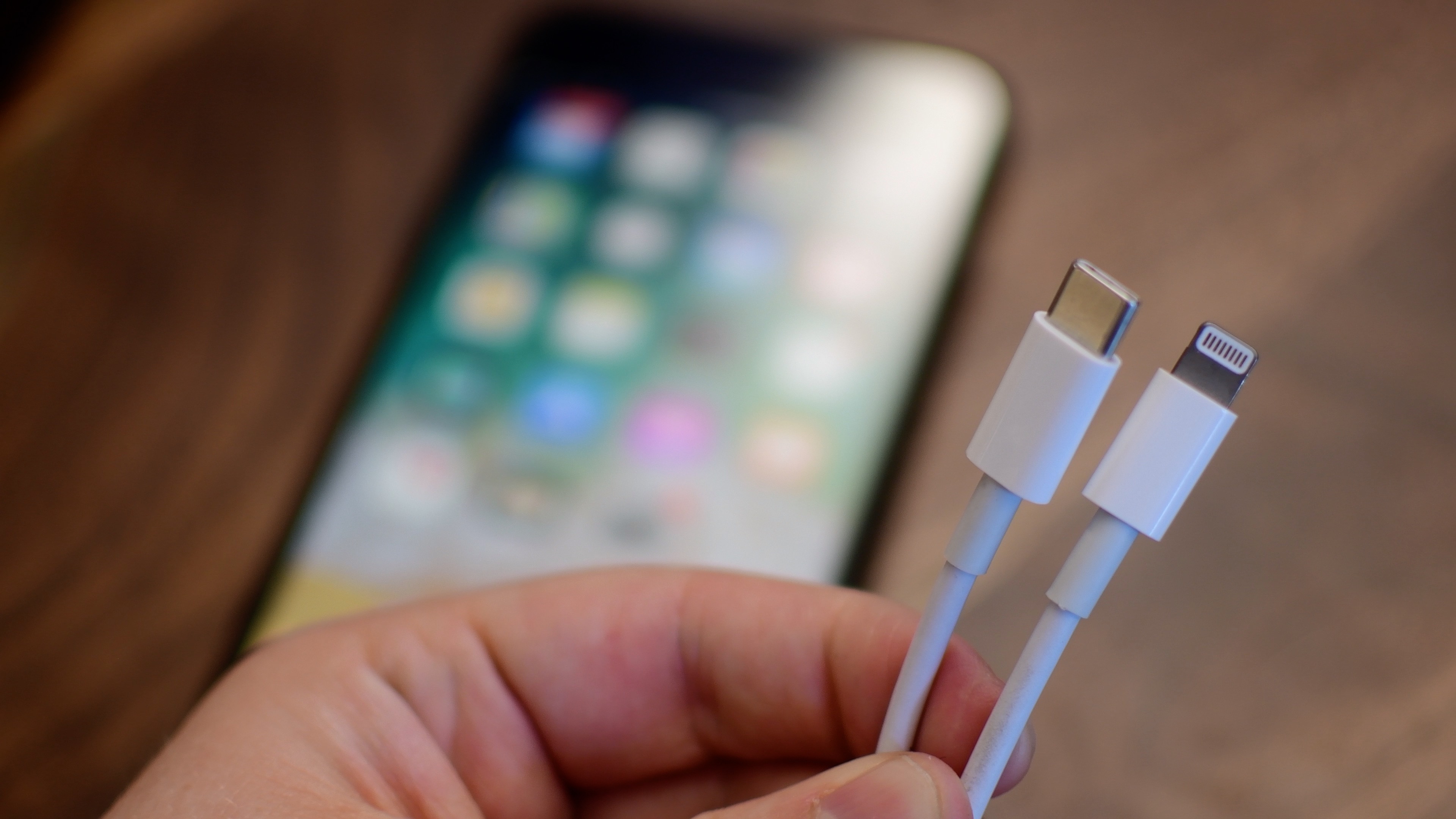 A photograph focused on a male hand holding both ends of Apple's Lightning to USB-C cable in the foreground, with an iPhone laid on a table blurred out in the background.