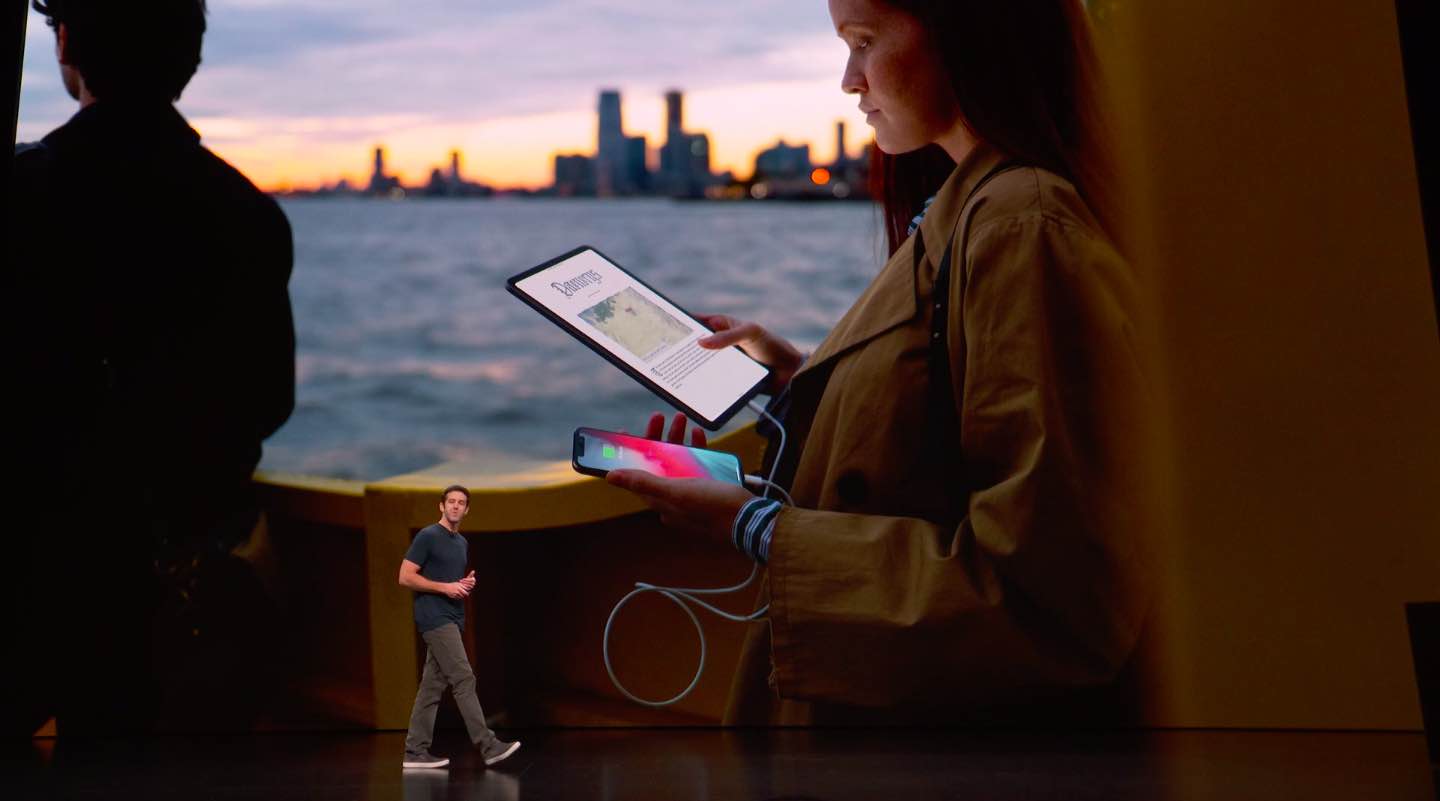 An image from Apple's 2018 event showing an executive walking in front of a slide displaying a woman holding an iPad Pro and an iPhone in her hands connected with a Lightning to USB-C cable