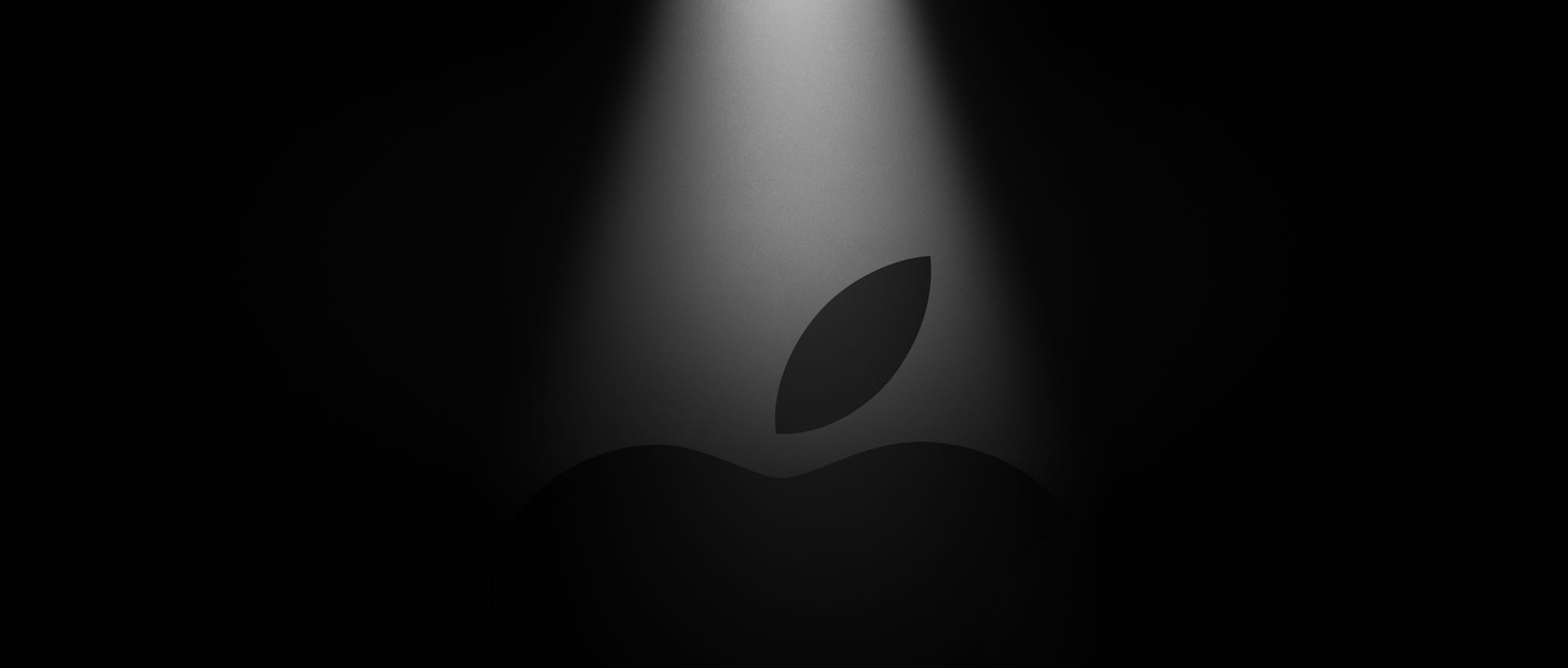 A teaser image showing the upper half of a black Apple logo, set against a dark background with a bright spotlight above
