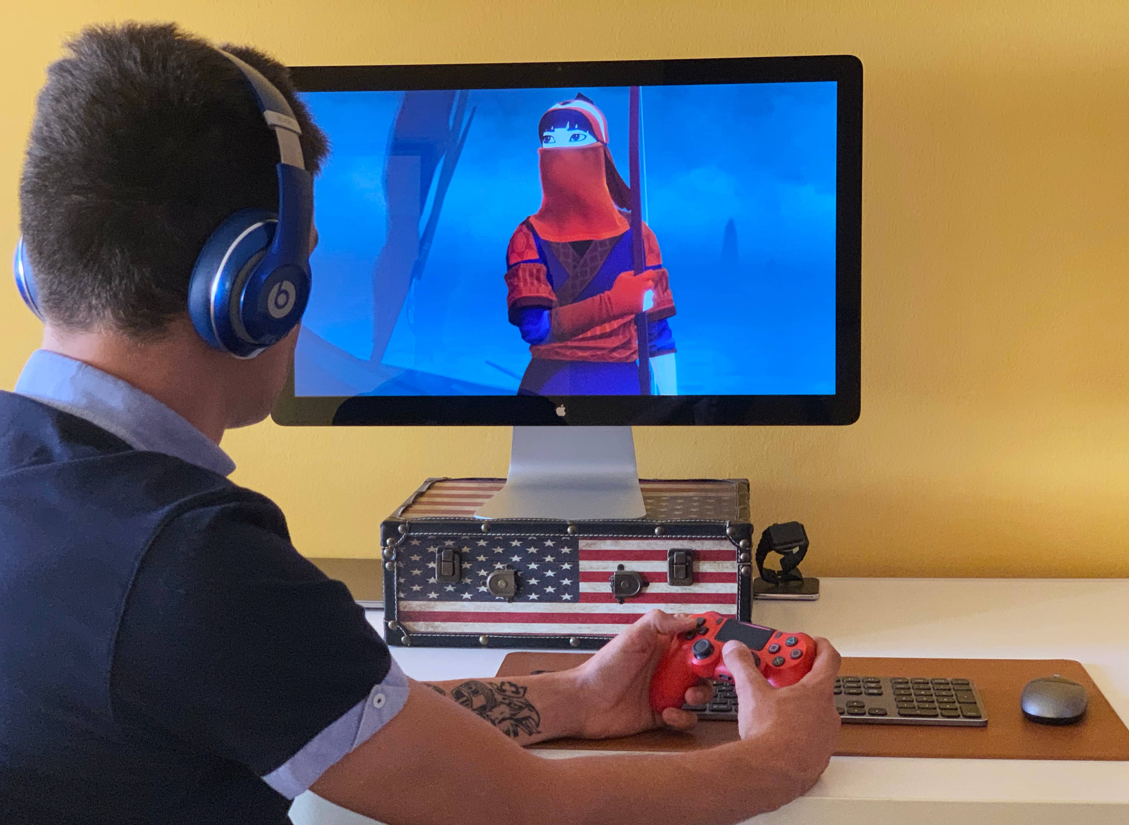 A photograph showing a young male sitting at their desk, holding a red PlayStation DualShock controller in their hand and playing an Apple Arcade game on their Apple Thunderbolt Display