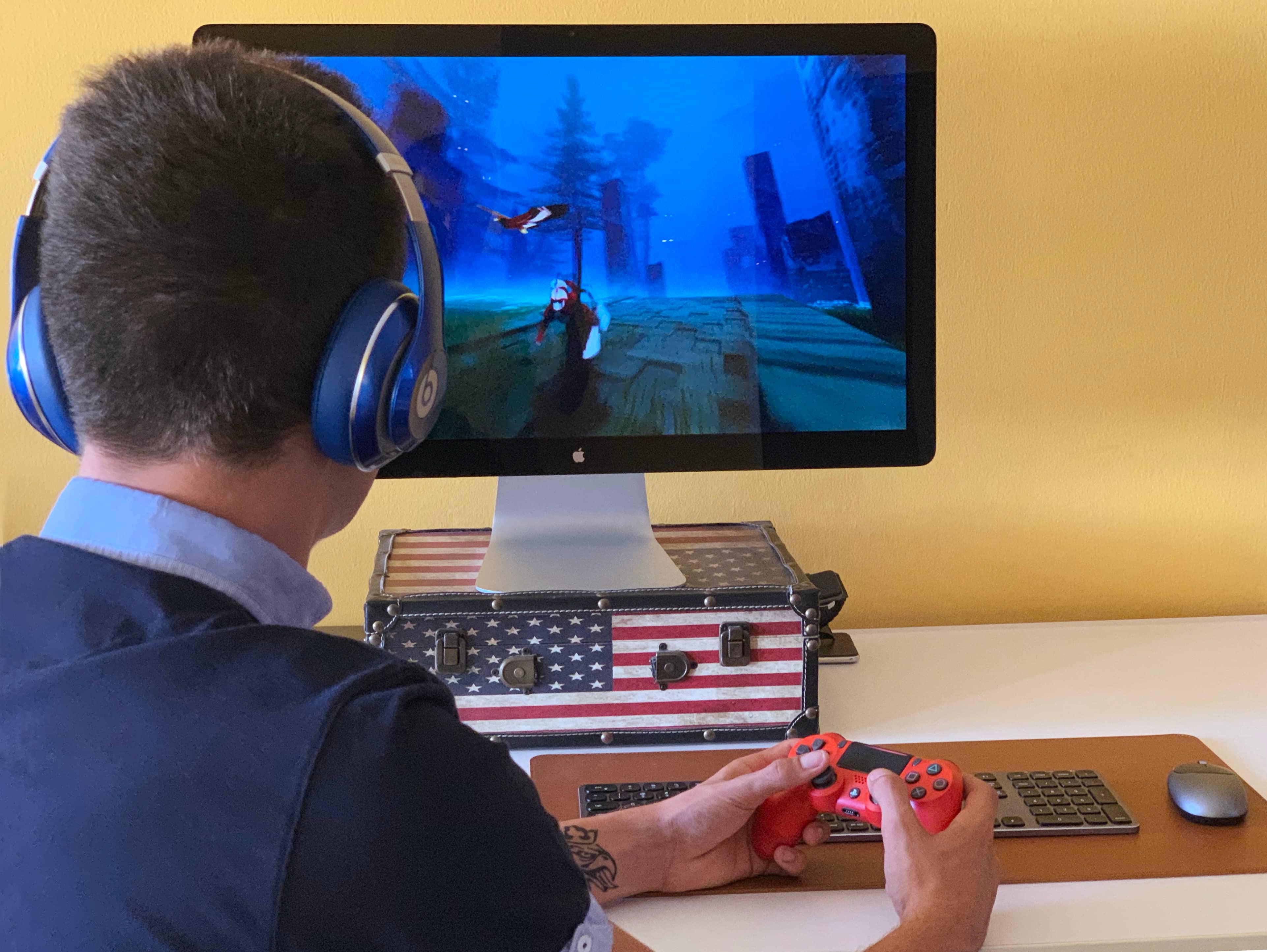 A photo showing a young male from the back who sits at a desk and holds a controller in his hands, playing a game on their Mac
