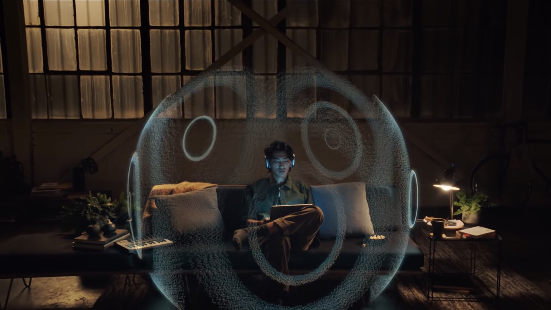 Apple illustration for spatial audio depicting a young male person sitting on their couch, wearing their AirPods Max and listening to music on iPad Pro