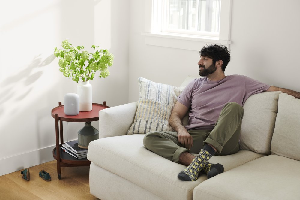 A marketing image showing a male sitting on a couch and listening to music on his Google Nest speaker