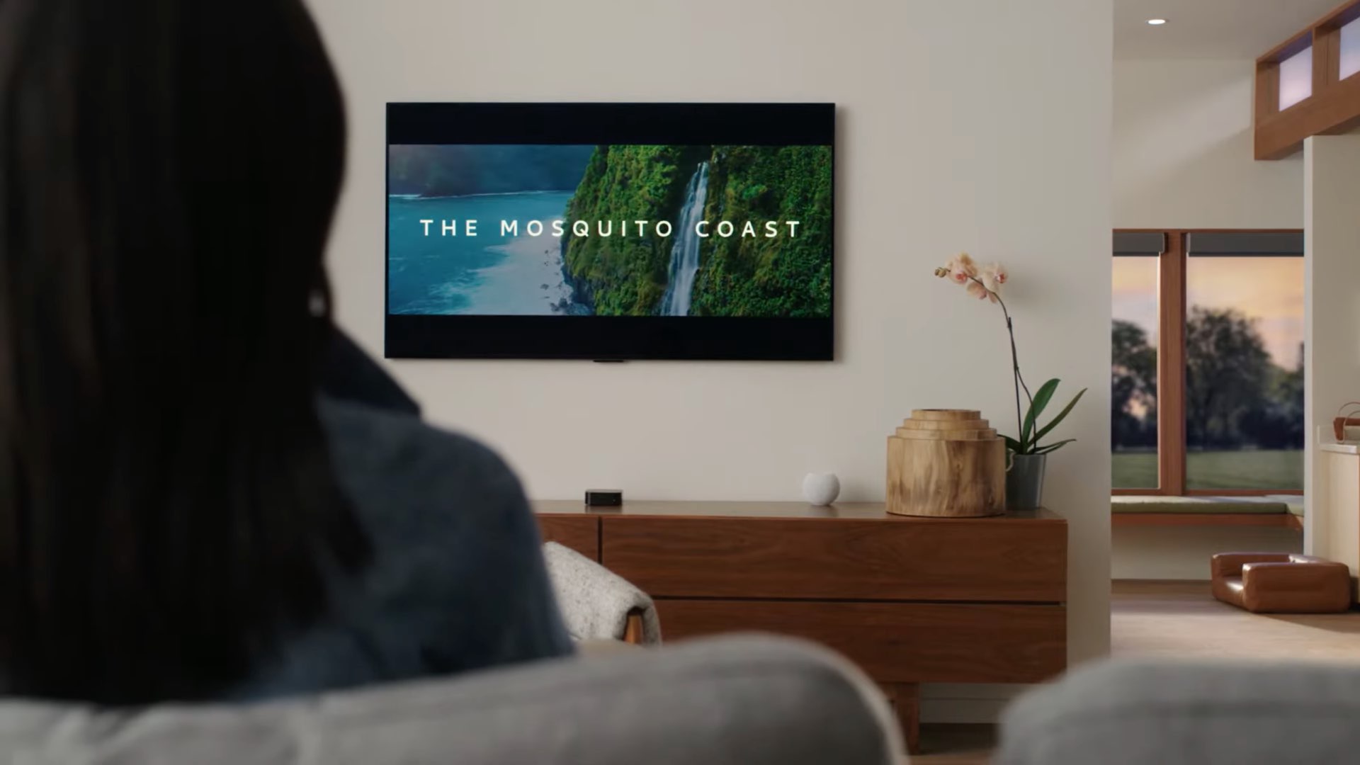 An image showing a person sitting on a couch and watching "Mosquito Coast" on their Apple TV with HomePod mini next to it