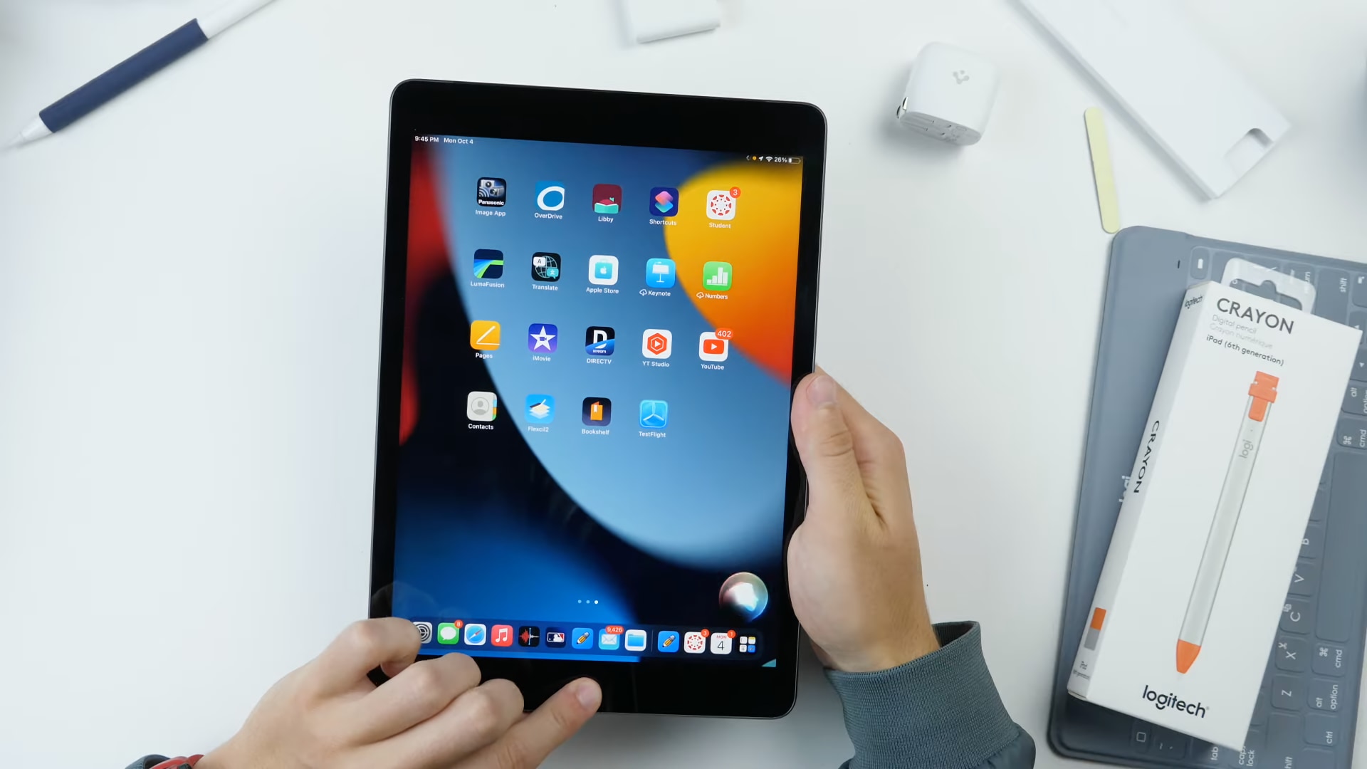 A top-down view of a white work desk with a your male's hands holding Apple's ninth-generation iPad in hands with the default home screen shown on the display