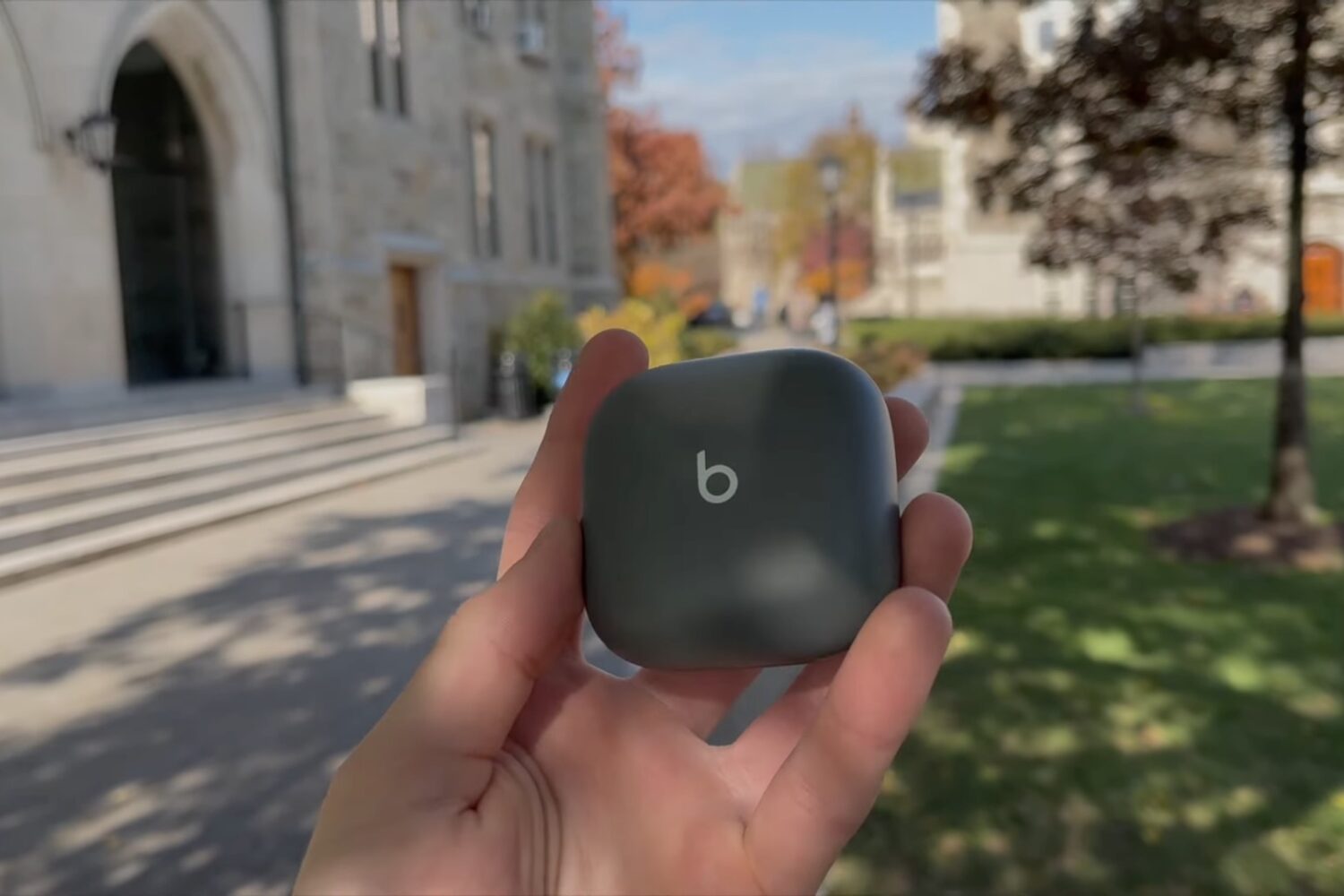 A young male's hand holding a closed wireless charging case for the Beats Fit Pro earbuds in a university park