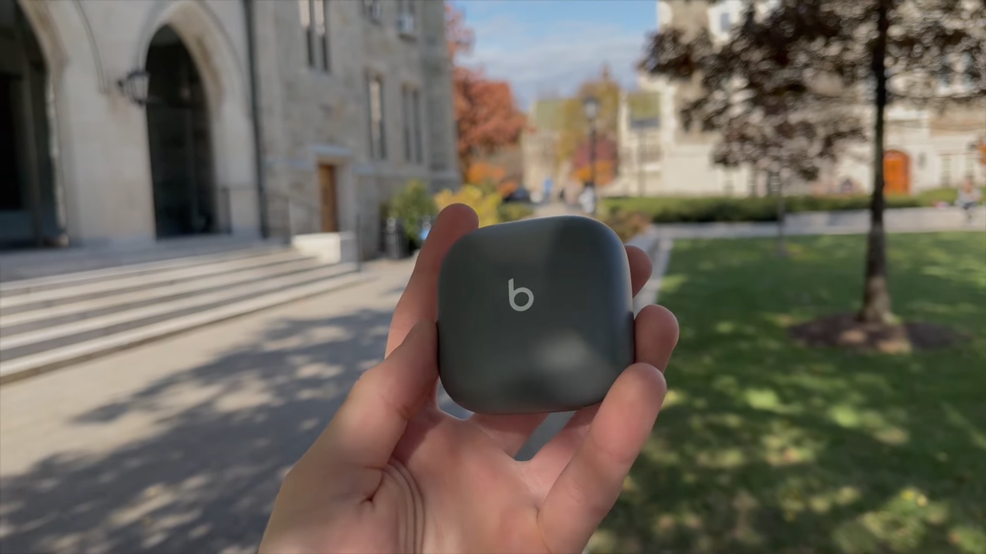 A young male's hand holding a closed wireless charging case for the Beats Fit Pro earbuds in a university park