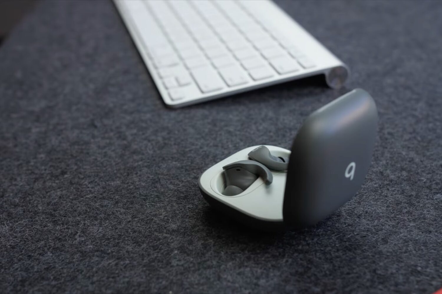 A Beast Fit Pro charging case with both earbuds shown with the lid open, laid flat on a gray work desk next to a white Apple wireless keyboard