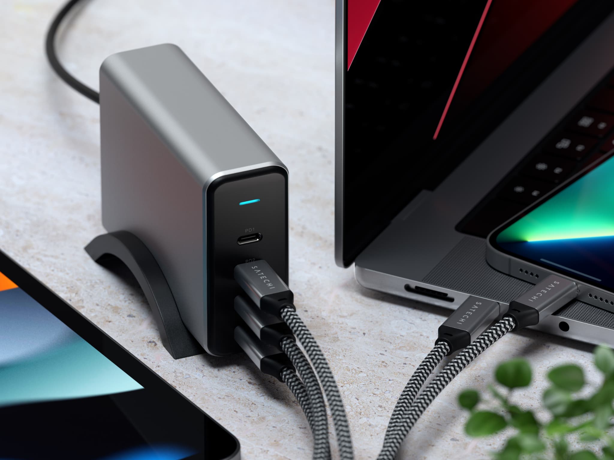 A lifestyle image showing a closeup of a 165W Satechi GaN charger on its stand, connected to two iPhones, iPad Pro and MacBook Pro
