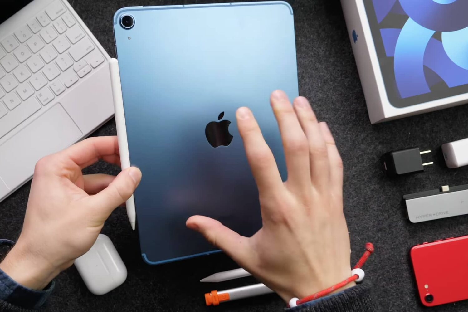 A top-down photograph showing a young male's left hand holding a blue iPad Air 5 with their right hand's fingers spread across the back of the tablet