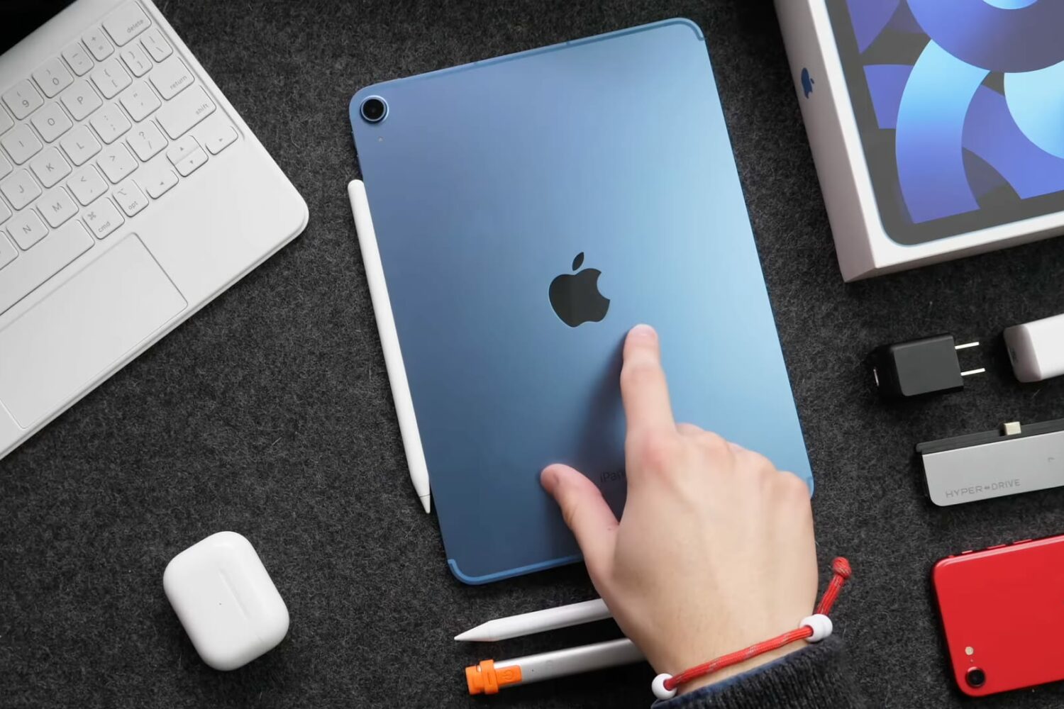 A top-down photograph showing a young male's right hand with the index finger pointing at a blue iPad Air 5 laid flat on a table facedown