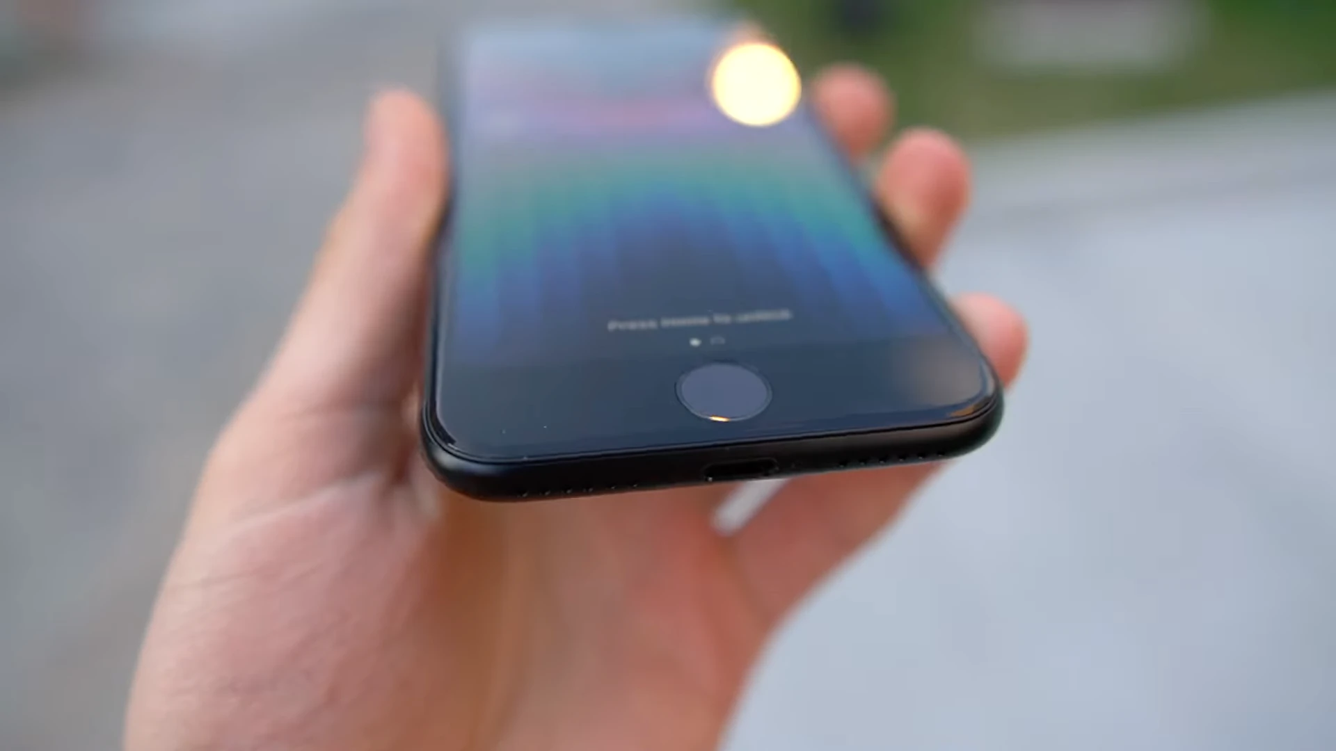 A top-down unboxing view showing a male's hands holding a black third-generation iPhone SE (model year 2022) with a focus on the Home button at the bottom