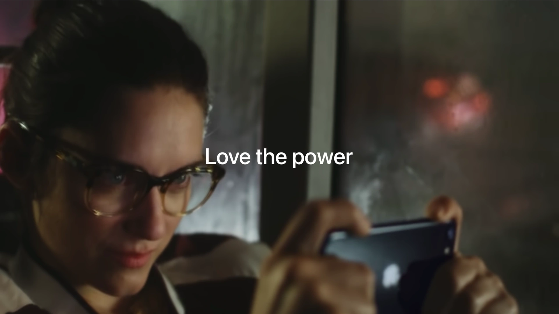 A still from Apple's video commercial showing a woman holding the third-generation iPhone SE in her hands in landscape orientation, looking intently at the screen, with the tagline "Love the Power" printed in white text