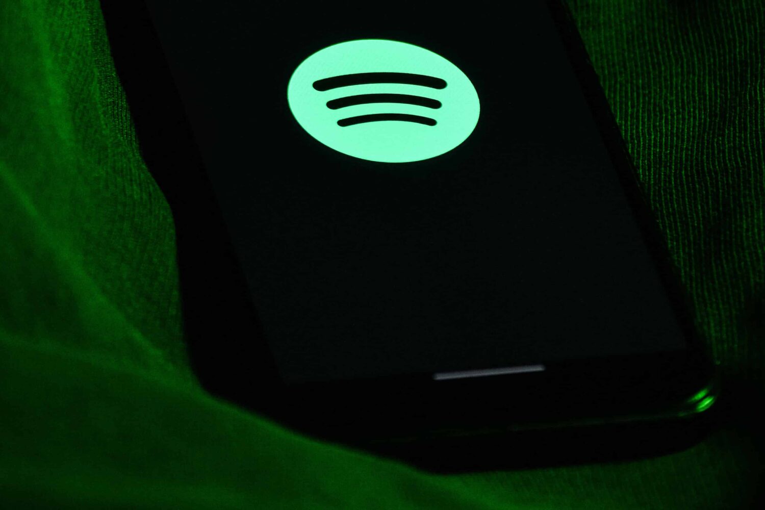 A photograph showing a back iPhone with a green Spotify logo displayed on the screen, laid on its back on a piece of green fabric