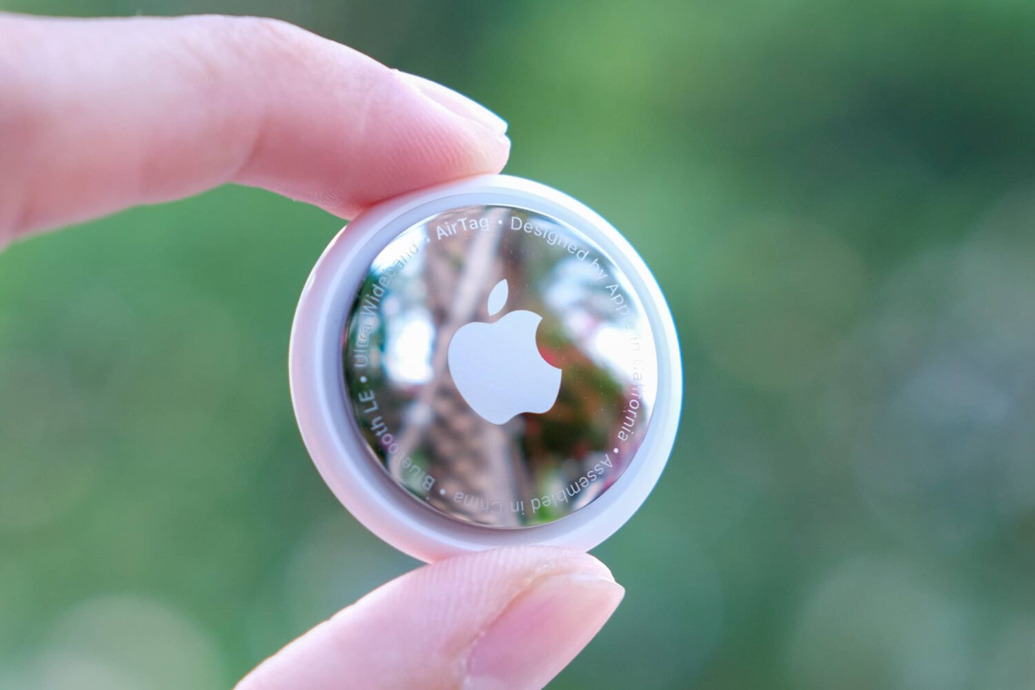 A closeup photograph of an Apple AirTag being held between a person's thumb and index finger, with the tracker's stainless steel back exposed to the camera