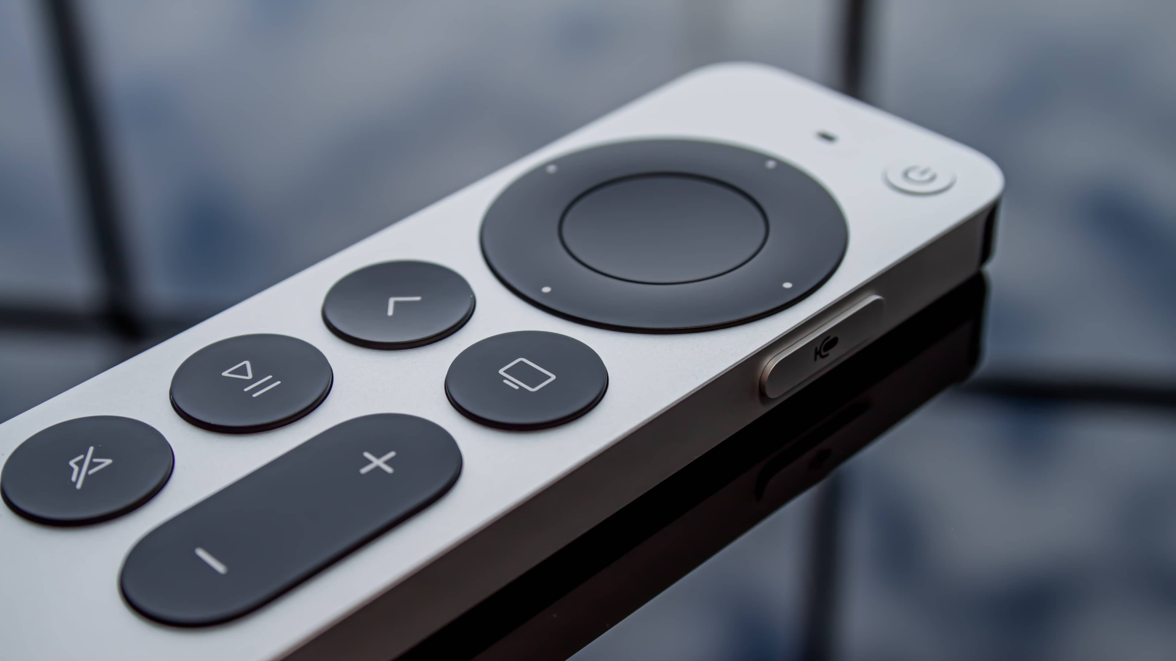 A redesigned Siri-powered remote for the Apple TV streaming box is shown laid flat on its back on a glass table in this close-up photograph.