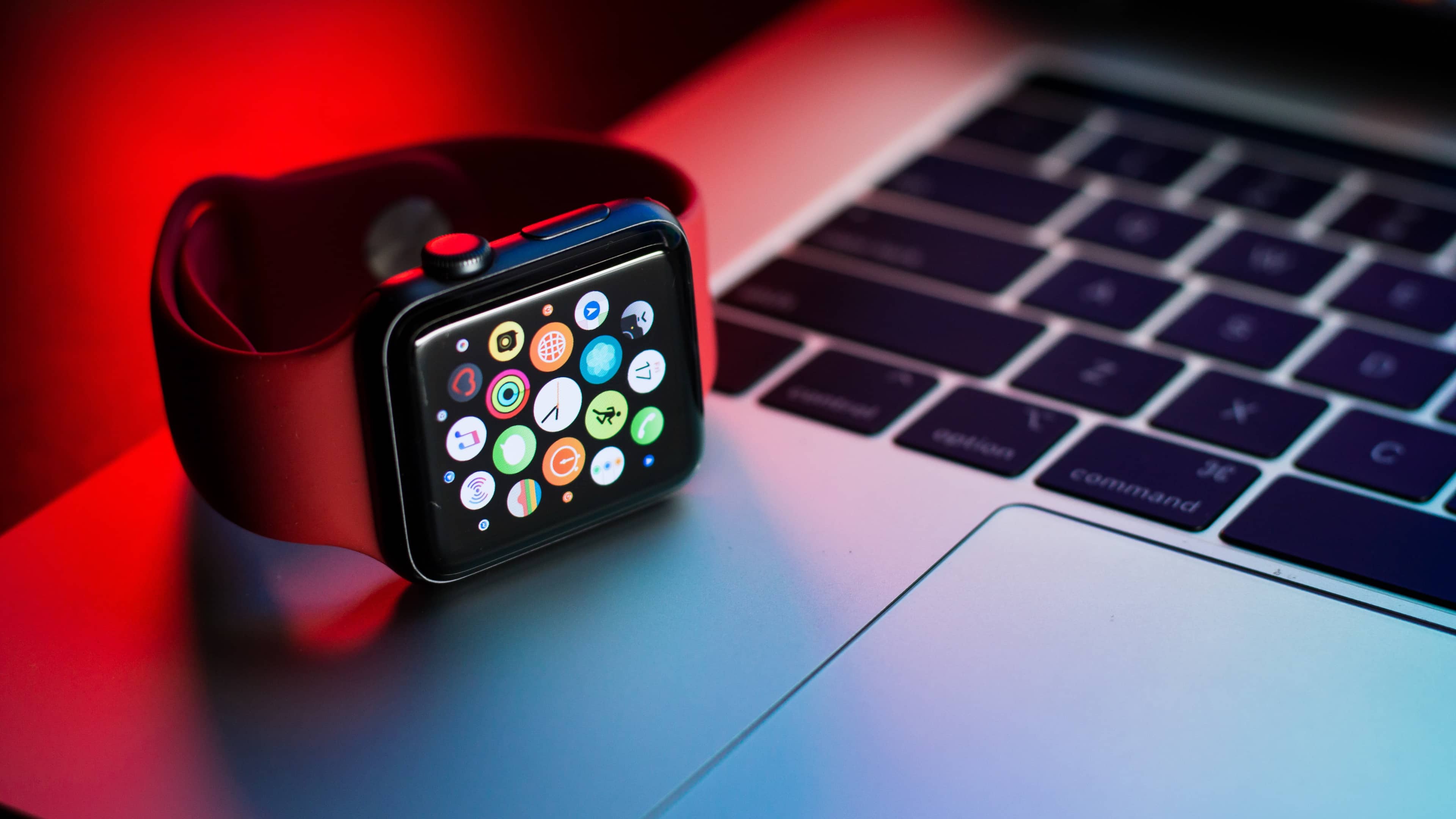 A black Apple Watch Series 7 is shown resting on its side next to the trackpad on Apple's MacBook Pro notebook in this lifestyle product photo