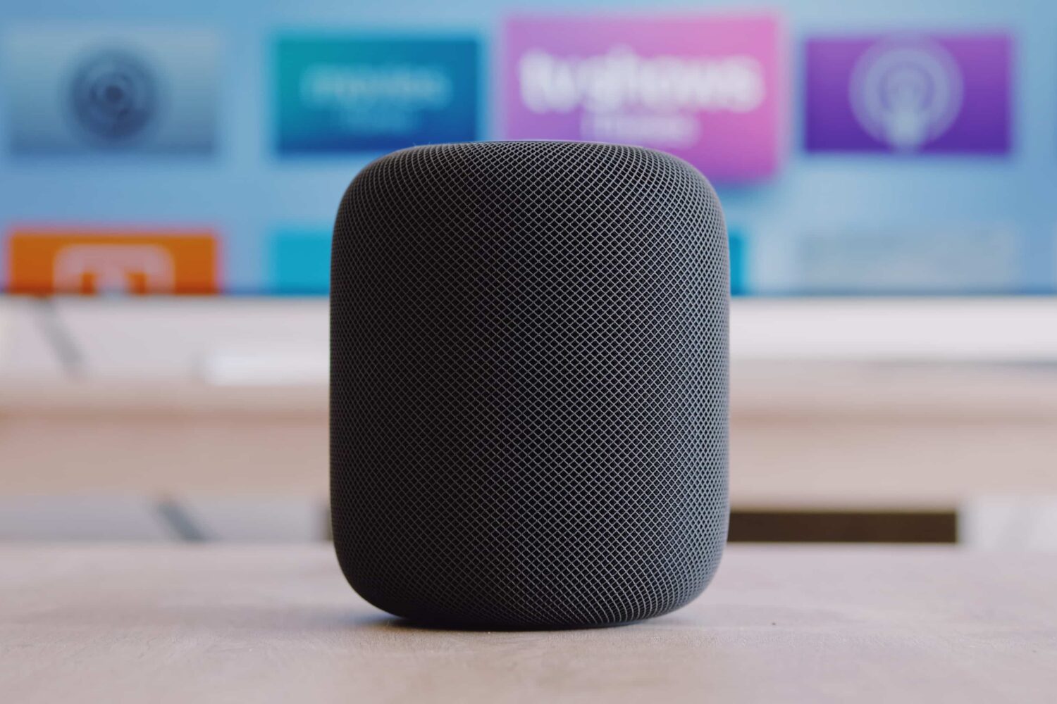 A black Apple HomePod wireless speaker laying on a table in front of a TV set which displays an Apple TV home screen