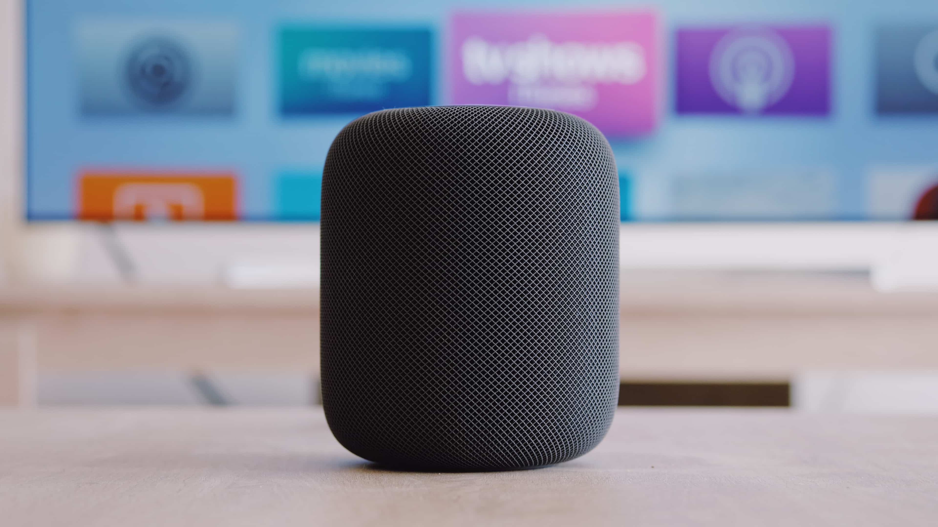A black Apple HomePod wireless speaker laying on a table in front of a TV set which displays an Apple TV home screen