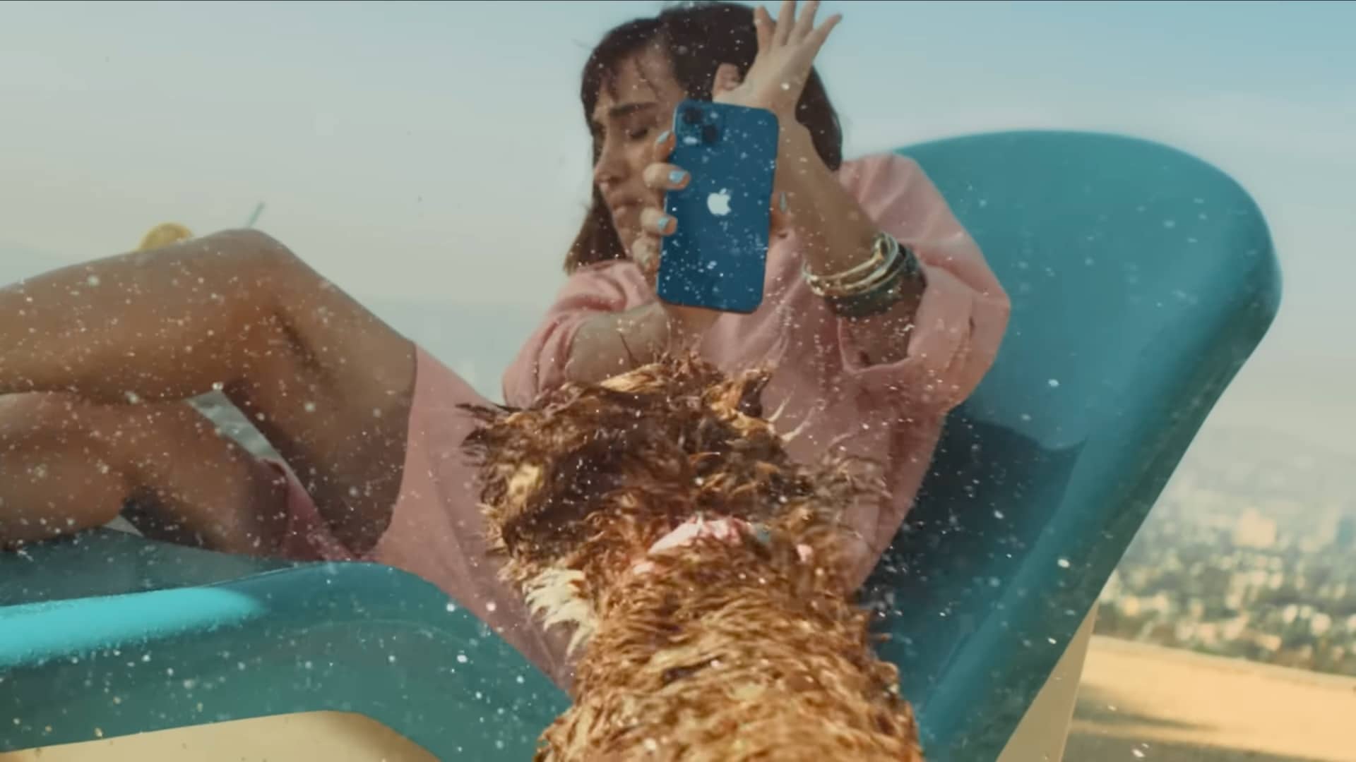 A scene from Apple's iPhone 13 ad, dubbed “Shake,” depicting a wet dog shaking to dry off next to a young woman sitting by the pool