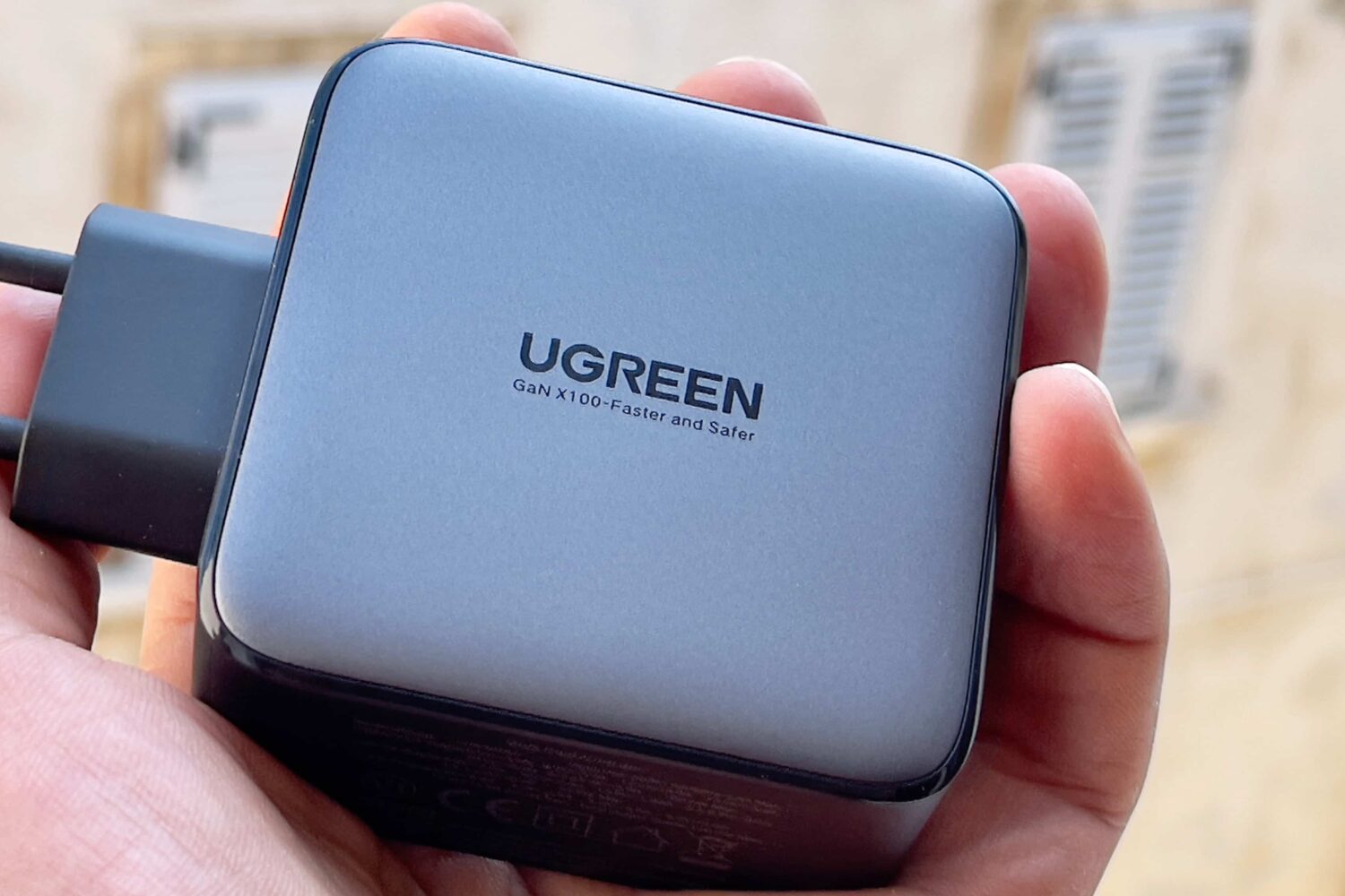 A male hand holding Ugreen's 100-watt Nexus power adapter, showing a Ugreen logo with the tagline "GaN X100-Faster and Safer" printed on the back of the casing