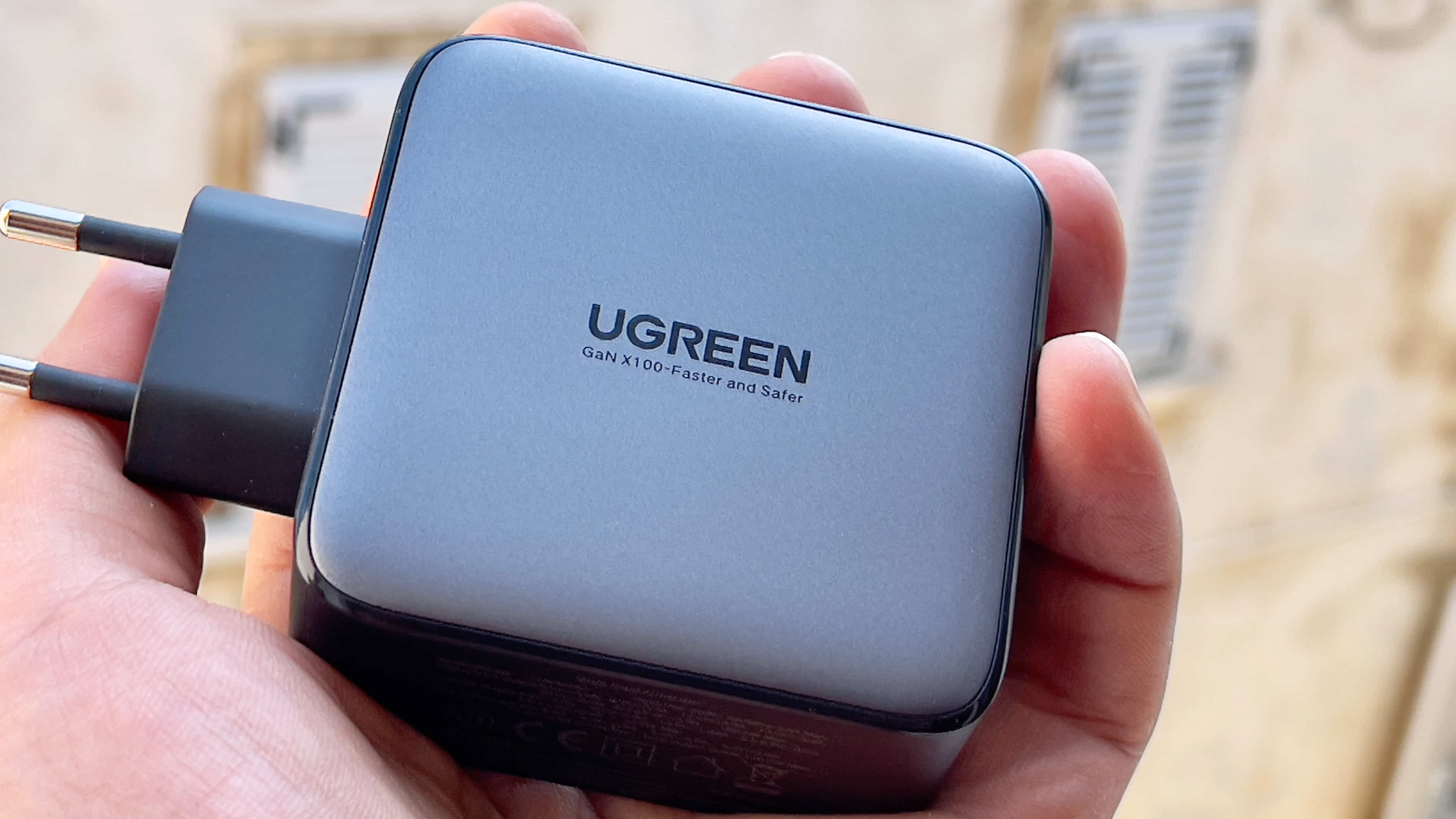 A male hand holding Ugreen's 100-watt Nexus power adapter, showing a Ugreen logo with the tagline "GaN X100-Faster and Safer" printed on the back of the casing