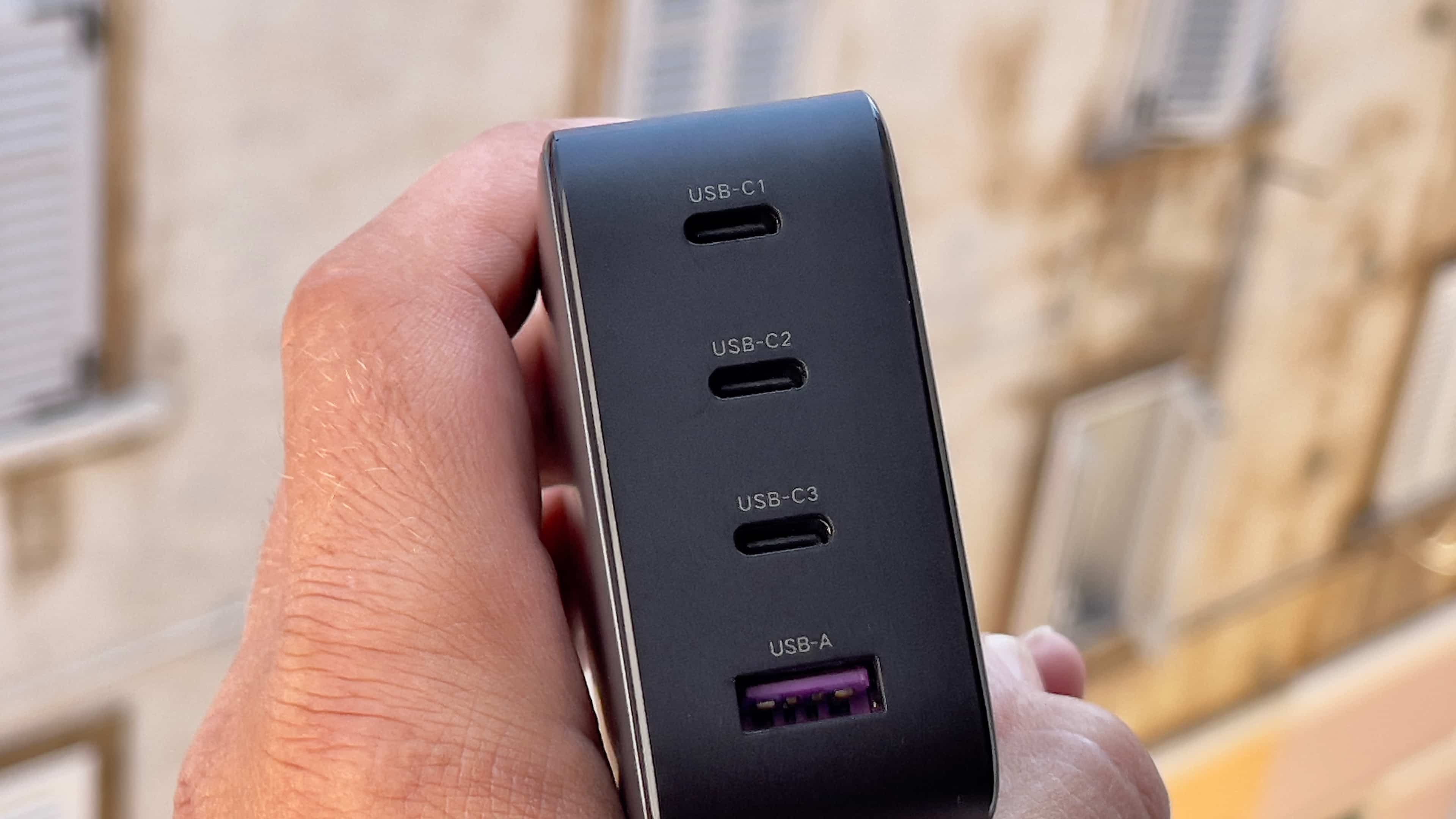 A male hand holding Ugreen's 100-watt Nexus power adapter, showcasing the charger's three USB-C ports and one USB-A port on the rear