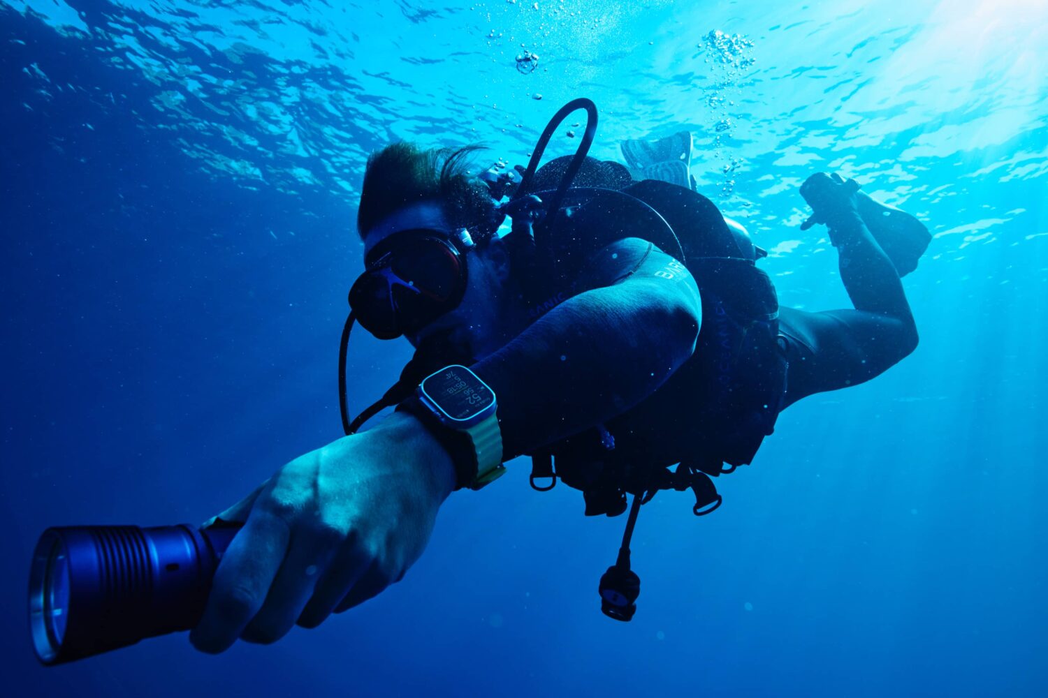 A scuba diver underwater using a flashlight and wearing Apple Watch Ultra with the Oceanic+ app providing dive computer functions