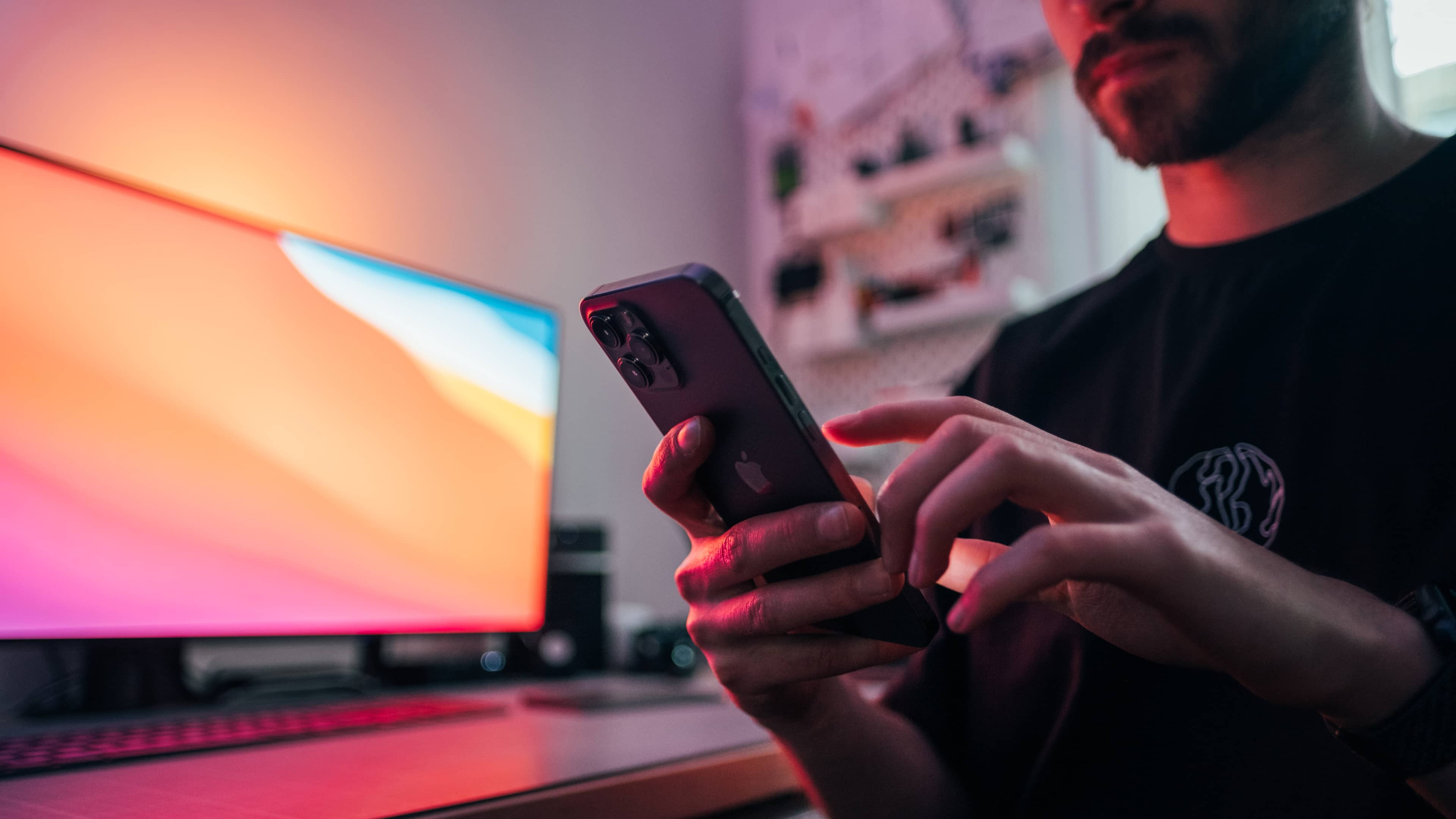 Ein junger Mann hält ein iPhone 12 Pro Max vor seinem Schreibtisch.  Im Hintergrund ist ein Monitor mit Big Sur-Hintergrundbild und einer raumgrauen Apple-Tastatur und einem Trackpad zu sehen