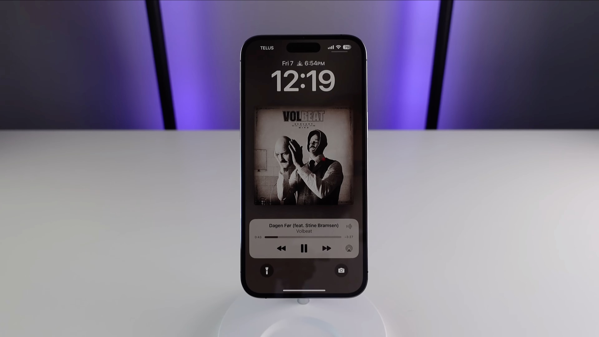 iPhone standing upright on a white table, showcasing fullscreen Now Playing controls on the lock screen