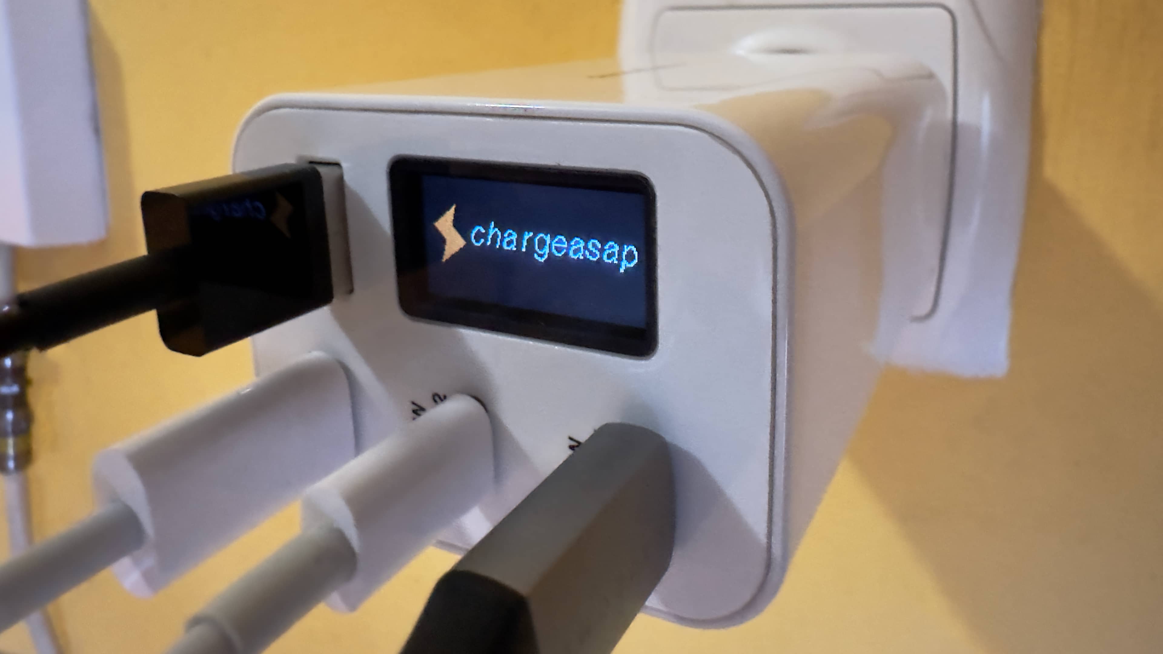 Closeup of Chargeasap's 150W Zeus power adapter plugged into a wall outlet, with cables in all of its USB ports and the OLED display showing branding logo