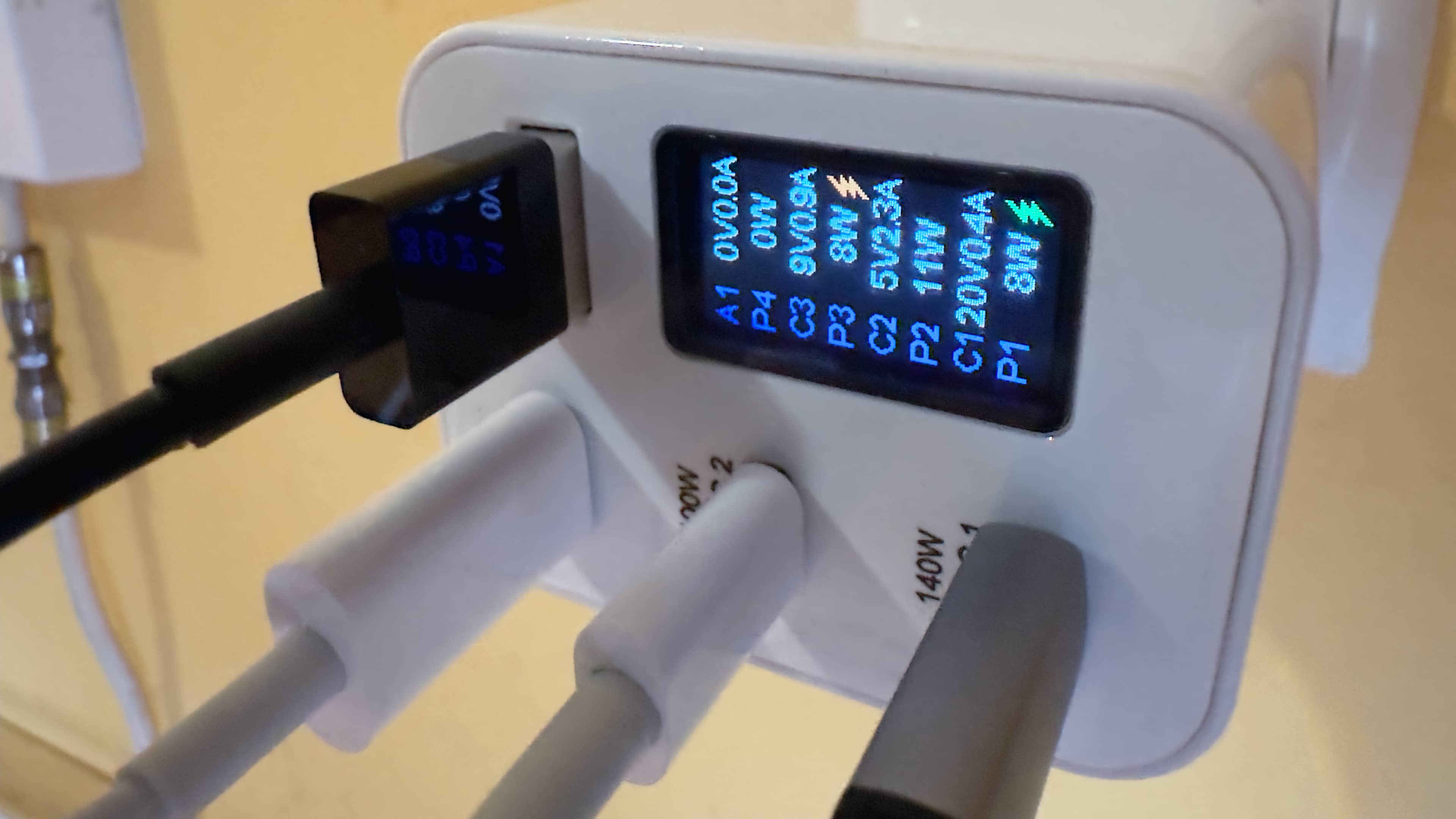 Closeup of Chargeasap's 150W Zeus power adapter plugged into a wall outlet, with cables in all of its USB ports and the OLED display showing voltage, amperage and wattage stats
