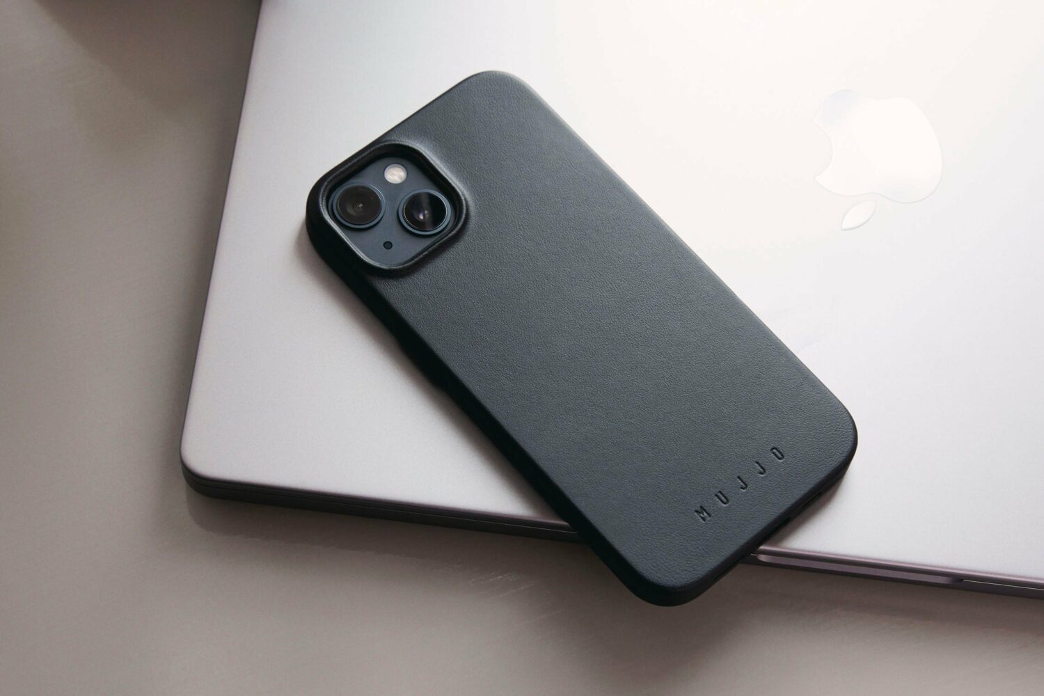 iPhone 14 in Mujjo's black leather case, set facedown on a Mac notebook