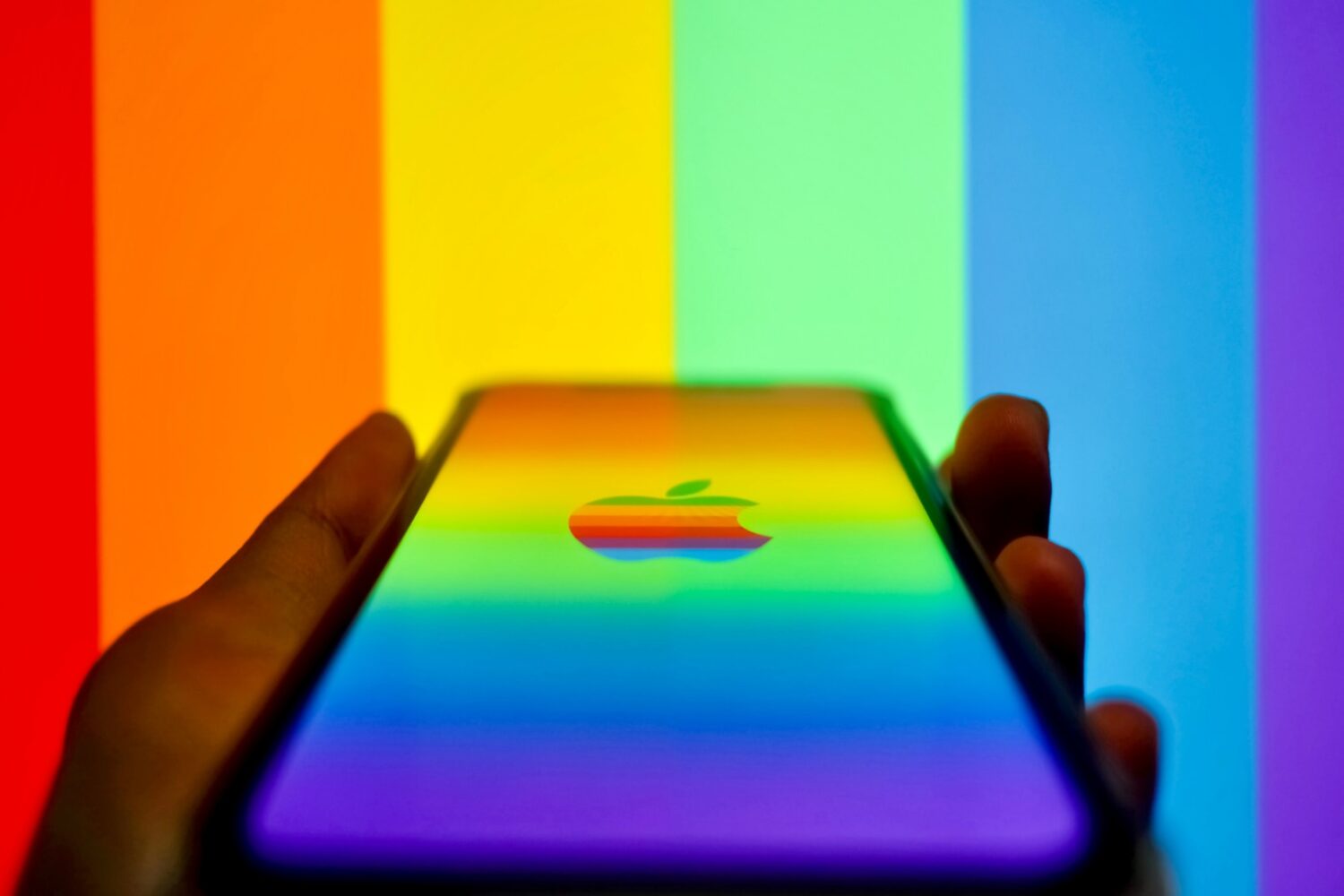 And holding iPhone displaying a rainbow Apple logo on the screen, set against rainbow stripes in the background