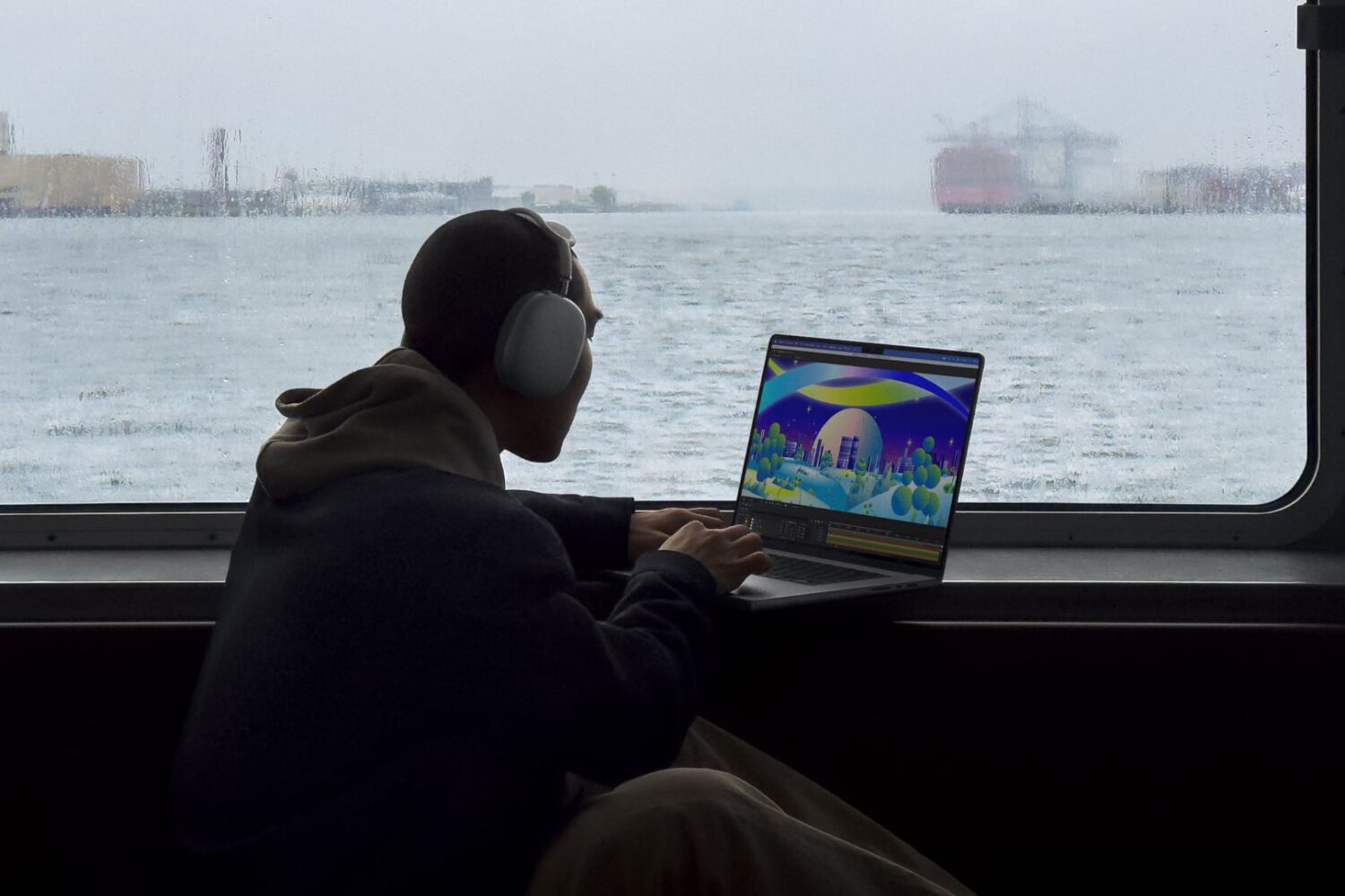 Young man on a boat on a rainy day, sitting next to a window and typing on his MacBook Pro