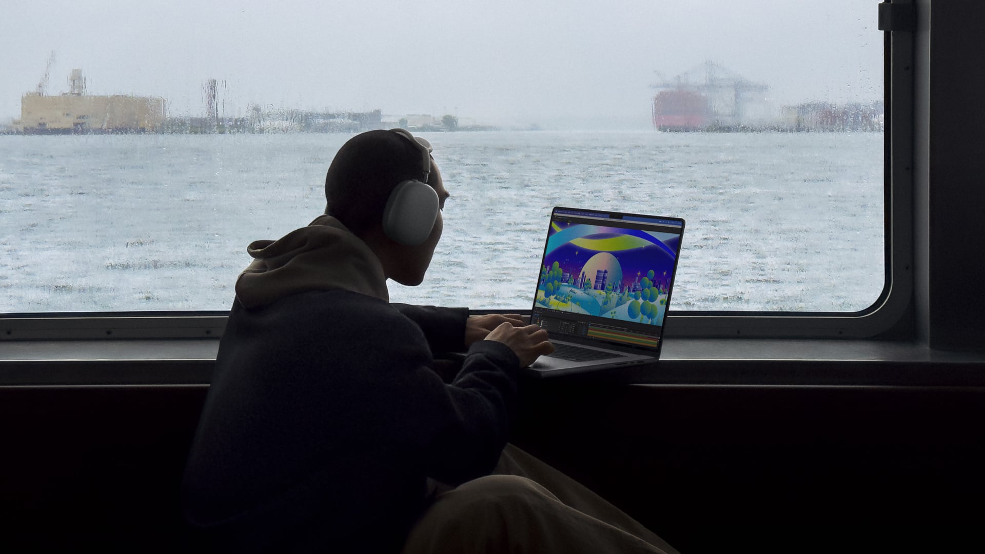 Young man on a boat on a rainy day, sitting next to a window and typing on his MacBook Pro