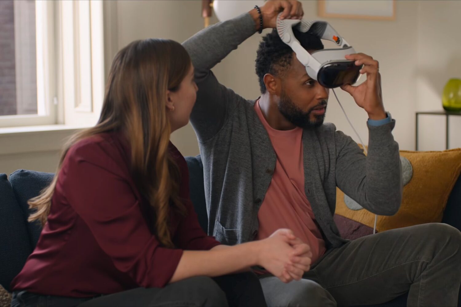 Man and woman sitting on a couch, with a man putting on Apple Vision Pro on his head
