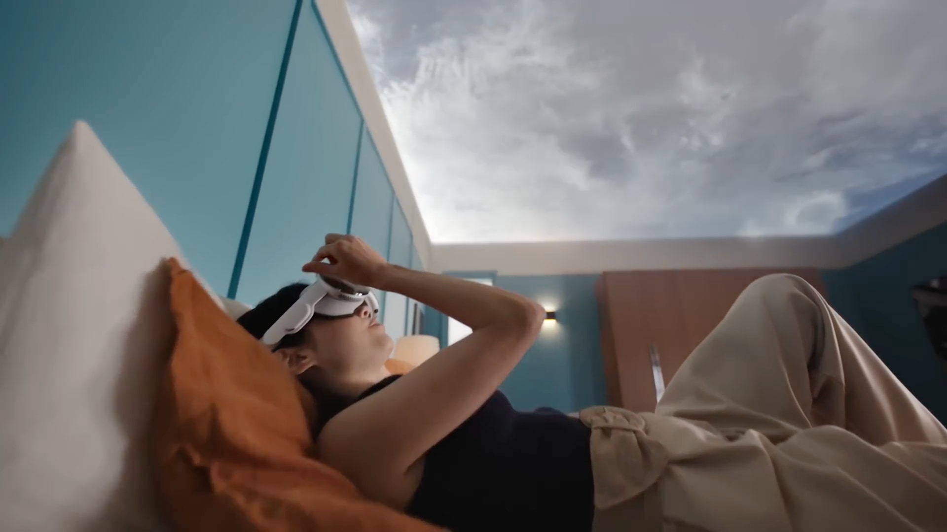 Young woman lying on bed while wearing Apple Vision Pro headset, with the view of her ceiling replaced by a virtual sky environment