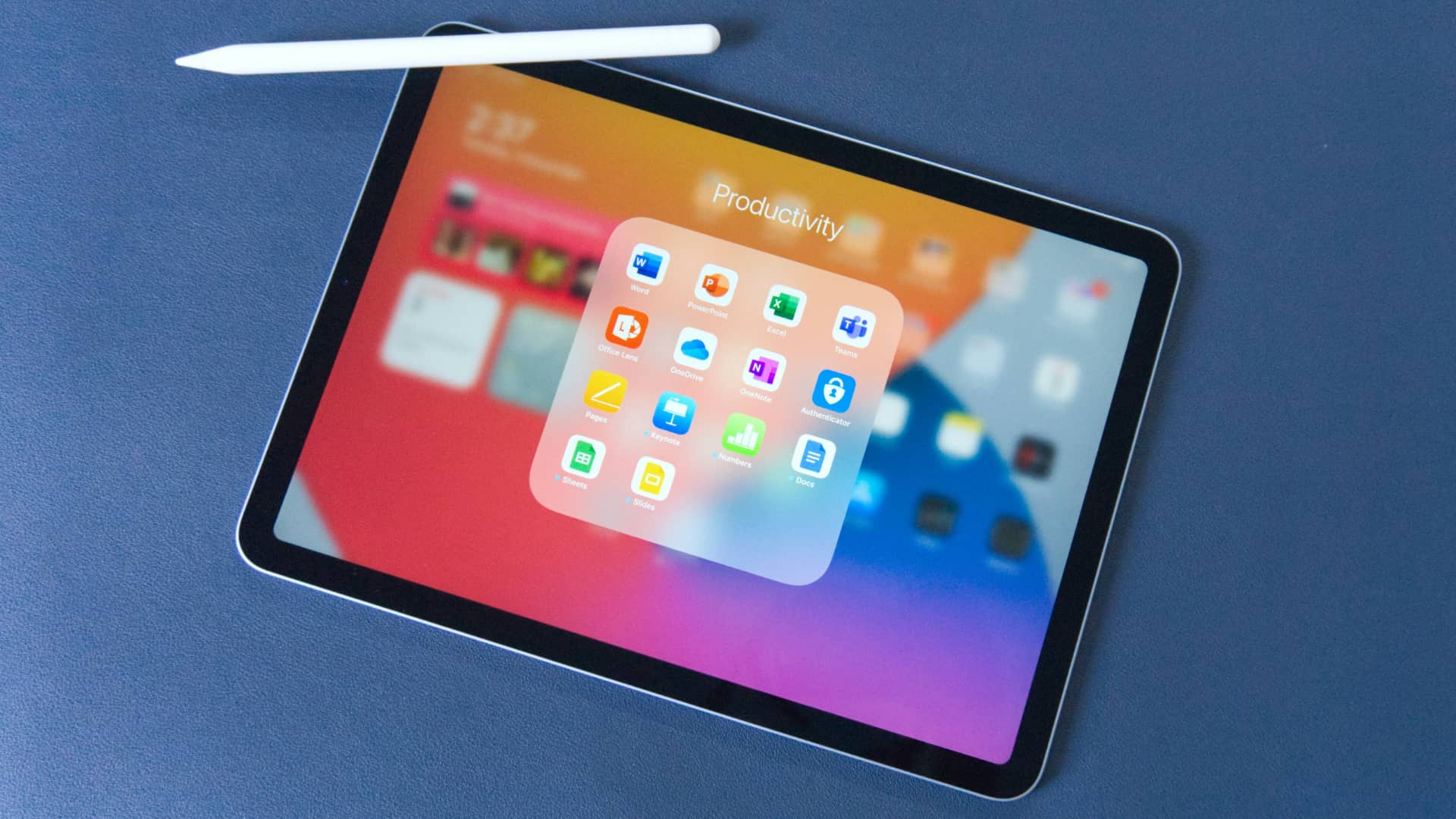 iPad Air laid on a blue leather desk pad, displaying a Productivity apps folder and having Apple Pencil magnetically attached along its edge