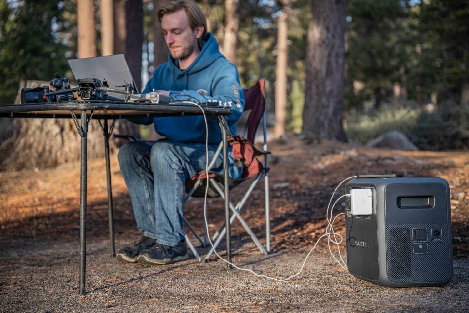 Man in the woods sitting at a wooden table and working on a computer, with his devices charging via Bluetti's AC180T portable power station