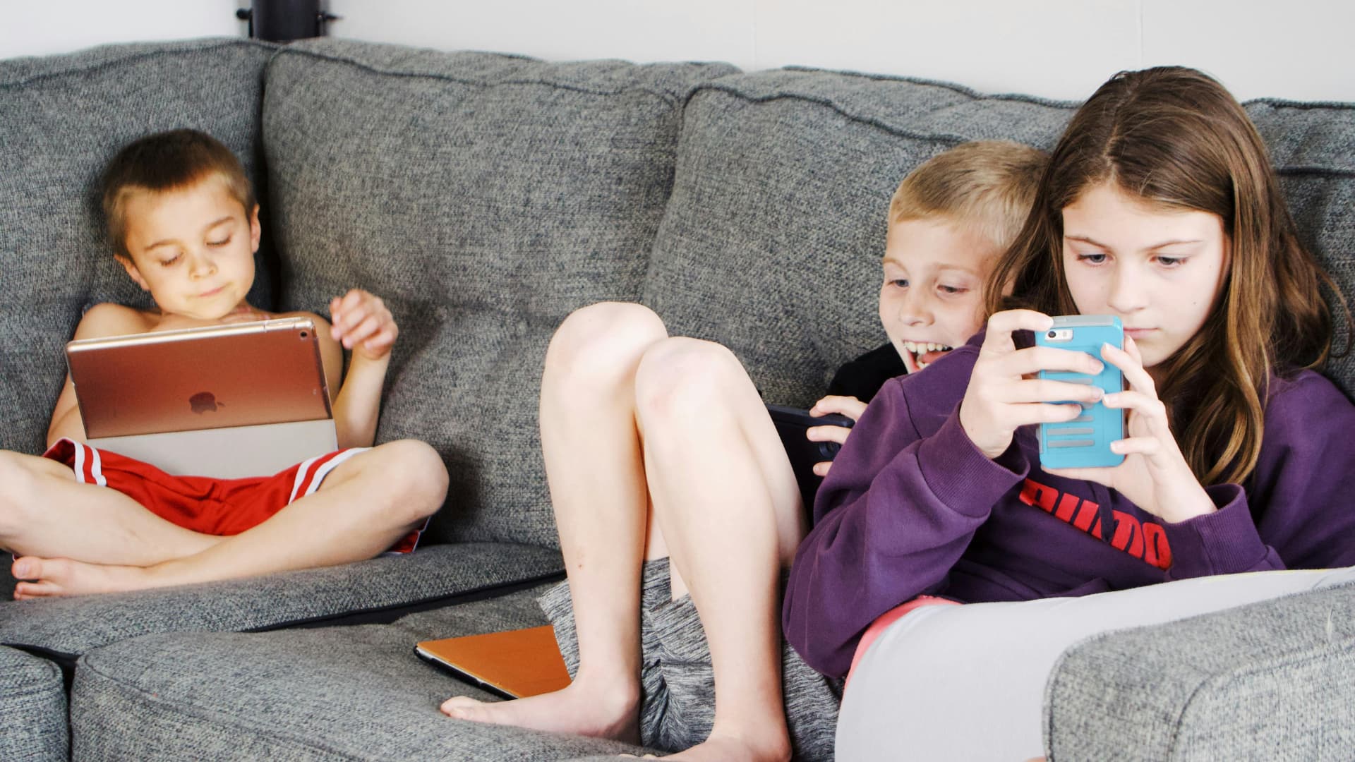 Three children sitting on a sofa and using iPhone and iPad